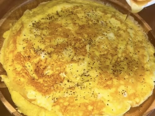 Fluffy yellow omelette sprinkled with black pepper in a bronze pan, liquid being poured over the top